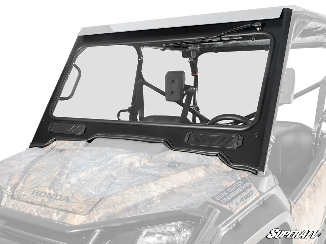 Close-up view of a SuperATV glass windshield mounted on a Honda Pioneer 1000 UTV. The windshield features a robust black frame with built-in sliding vents and a manual wiper, showcasing intricate details like the angular design of the vents and the secure mounting system. The hood of the UTV displays a camo pattern with the Honda logo.
