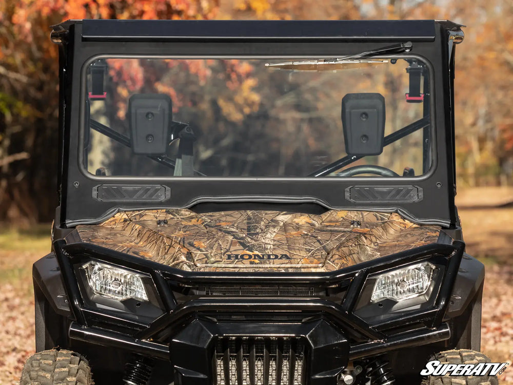 Front view of a black Honda Pioneer 1000 UTV equipped with a SuperATV glass windshield, framed by an autumn backdrop with colorful trees. The windshield features a clear view, sliding vents, and a manual wiper, set against the UTV's camouflage hood and rugged design, highlighting both functionality and style.
