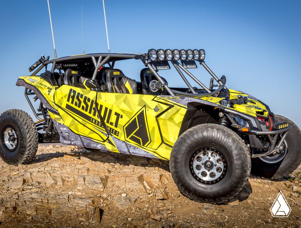 Customized yellow and black Can-Am Maverick X3 MAX UTV with Assault Industries graphics and upgrades, equipped with LED light bar and off-road tires, showcased on rocky terrain.