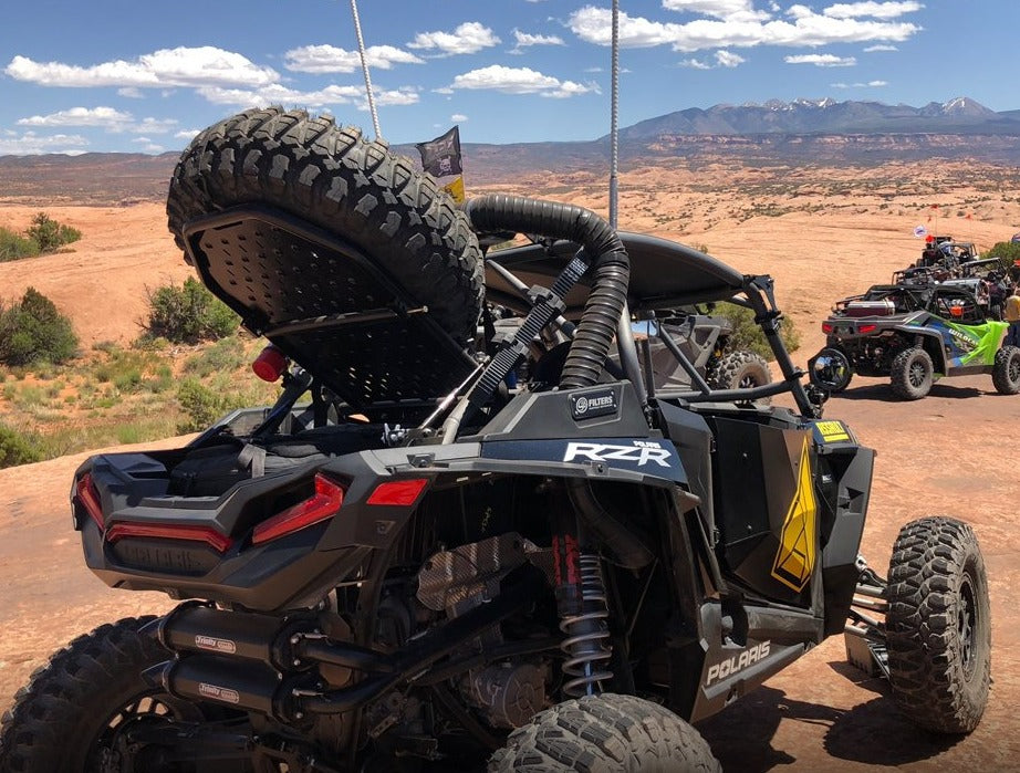 Assault Industries Adventure Rack mounted on a Polaris RZR, carrying a spare tire, with desert landscape and other off-road vehicles in the background.