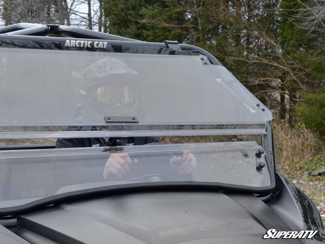 A rider in a helmet is visible through the clear, scratch-resistant flip windshield of an Arctic Cat Wildcat Trail utility vehicle. The windshield is securely mounted, showing parts of its adjustable latch and sturdy frame, set against a backdrop of trees and cloudy skies.