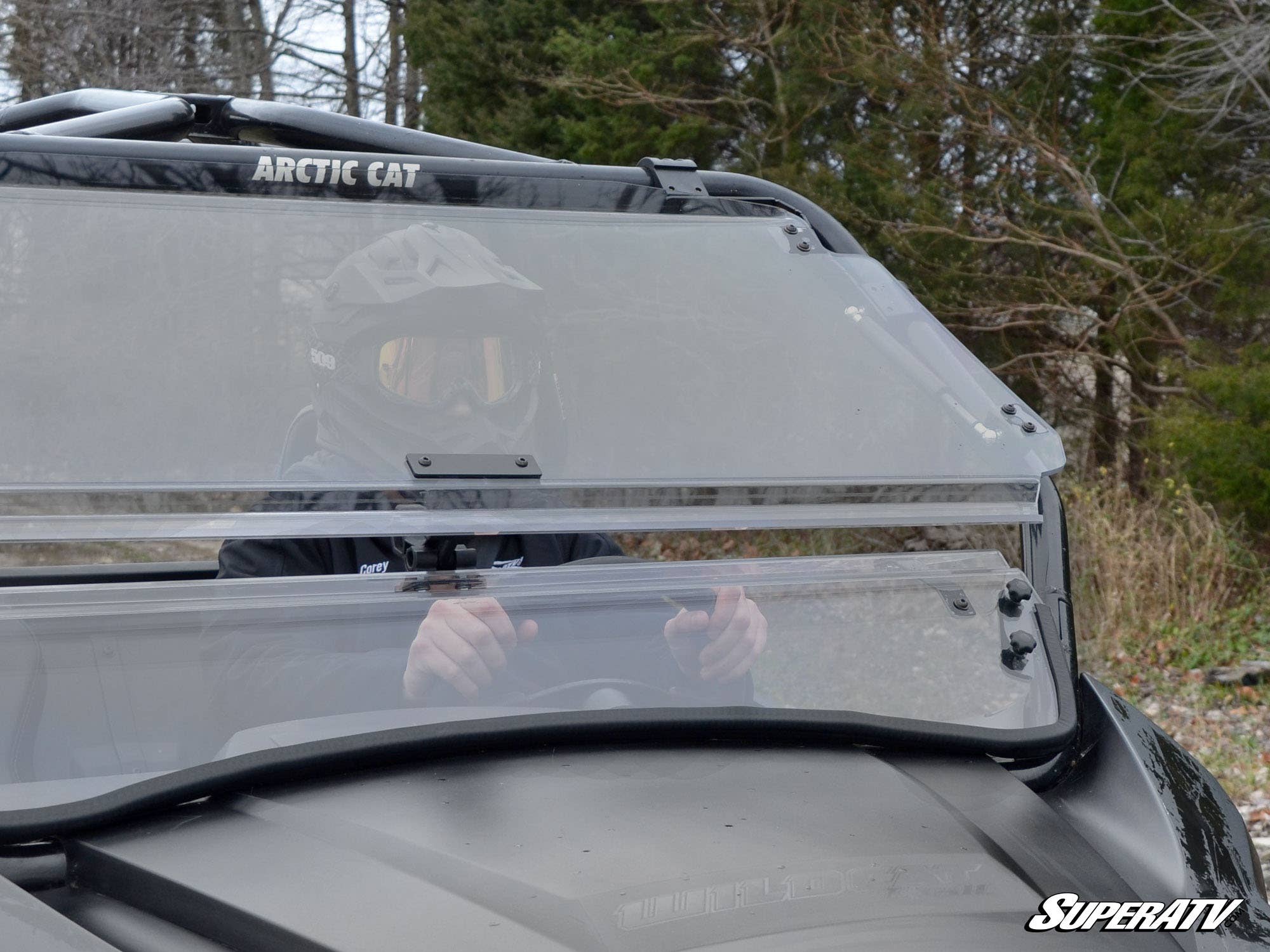 A rider in a helmet is visible through the clear, scratch-resistant flip windshield of an Arctic Cat Wildcat Trail utility vehicle. The windshield is securely mounted, showing parts of its adjustable latch and sturdy frame, set against a backdrop of trees and cloudy skies.